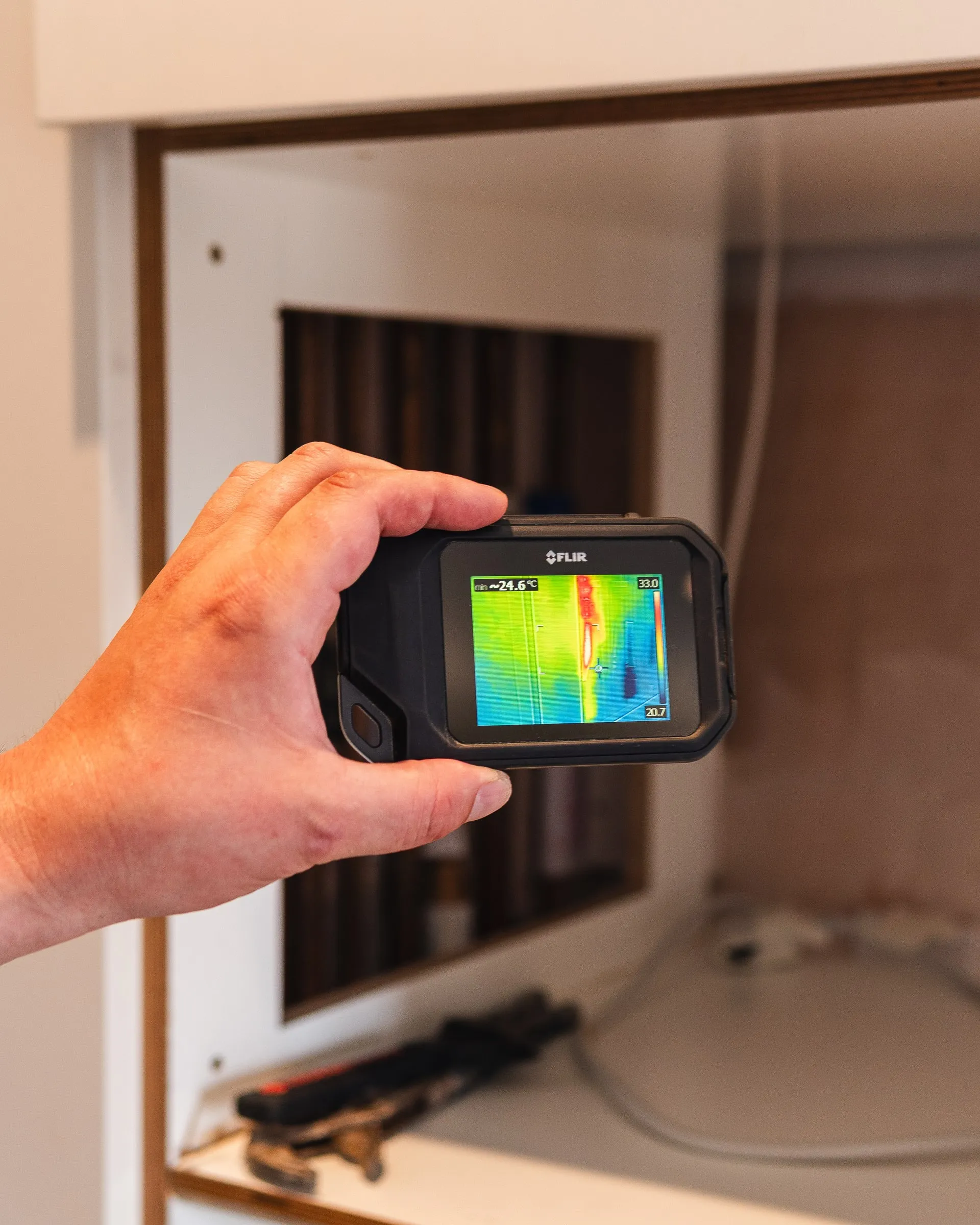 Engineer's hand holding a thermal camera up to an open service cupboard, the screen showing the warm pipe behind the panel